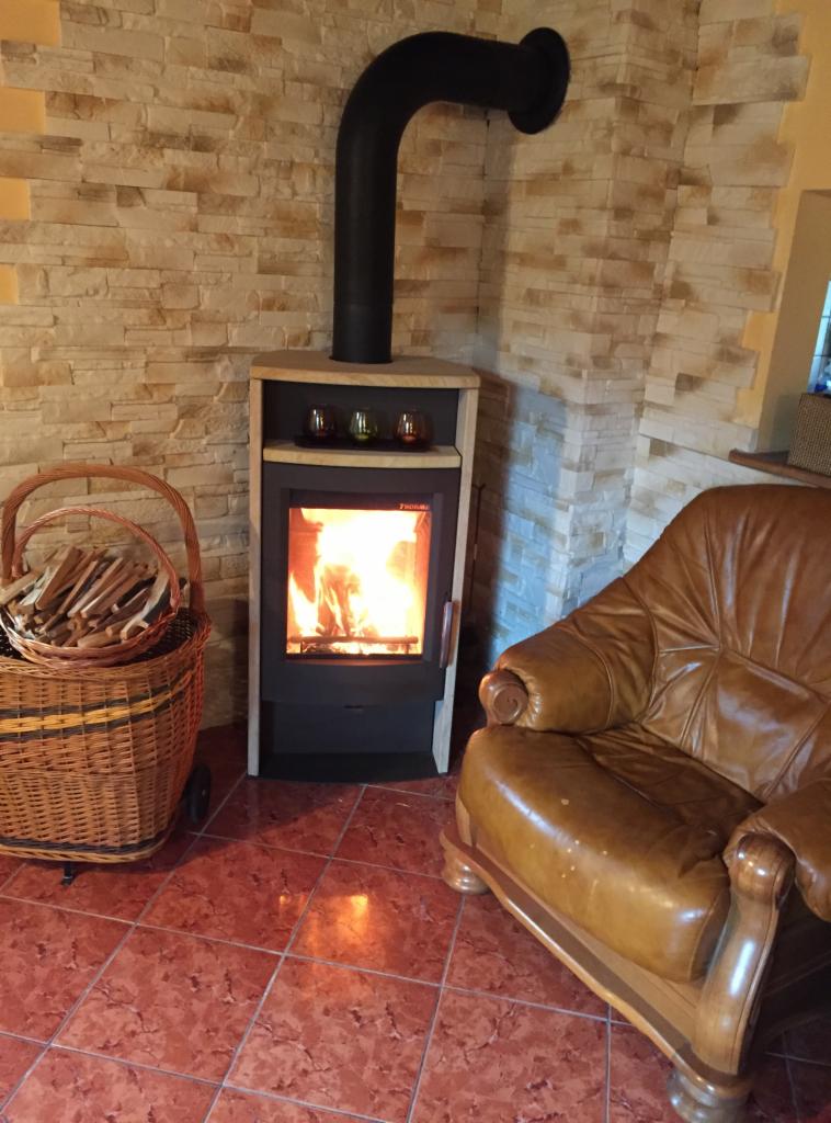 Nowoczesny piec na paliwo stałe z czarnym kominem, palącym się ogniem w palenisku, dekoracyjnymi szklankami na górnej półce, koszem z drewnem obok i skórzanym fotelem w tle, na tle ściany...