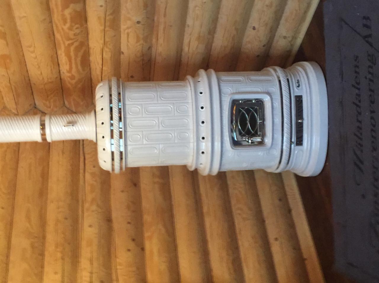 Biały piec kaflowy z ozdobnym wzorem cegiełek i przeszklonymi drzwiczkami paleniska, podłączony do białego komina, na tle drewnianej ściany z bali.