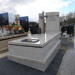 ZAKO Zaczernie - Jasny nagrobek z krzyżem i zniczem na cmentarzu w Zaczerniu. Kamienna konstrukcja z widoczną fakturą, otoczona innymi grobami i drzewami.