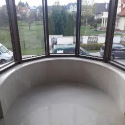 Półokrągłe wnętrze z dużymi oknami i marmurowym wykończeniem ścian oraz podłogi z jasnych płytek, widok na posesję z zaparkowanymi samochodami.