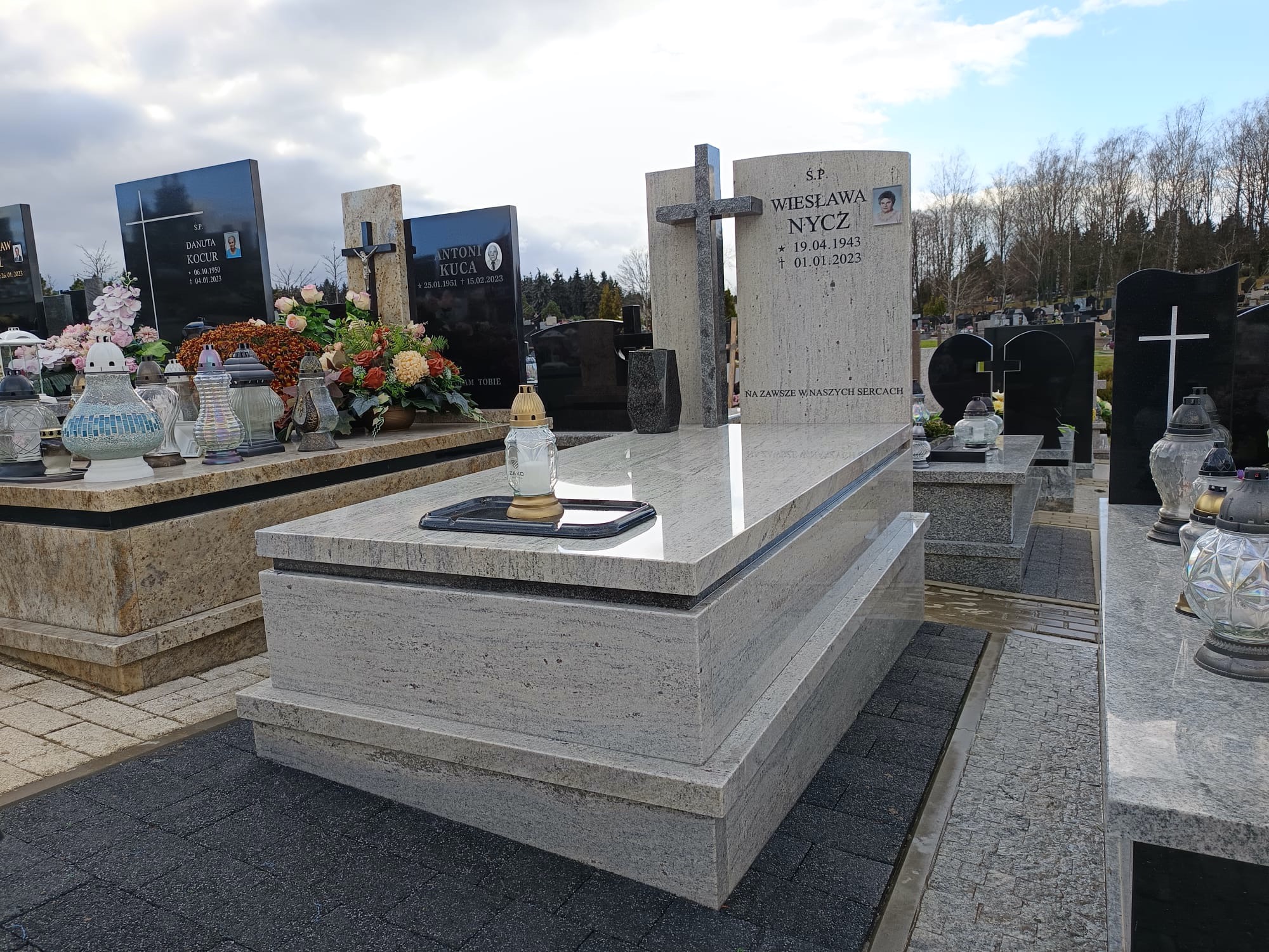 Jasny nagrobek z krzyżem i zniczem na cmentarzu w Zaczerniu. Kamienna konstrukcja z widoczną fakturą, otoczona innymi grobami i drzewami.