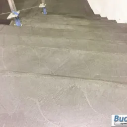 Szare schody z żywicy z widoczną fakturą i metalową balustradą w trakcie montażu, zabezpieczoną niebieską taśmą.