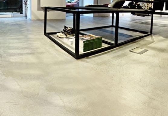 Jasnoszara, gładka posadzka z widoczną teksturą, z czarną, metalową konstrukcją meblową w tle, na której leżą buty i książki.