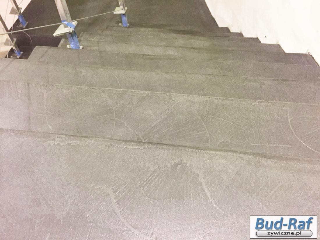 Szare schody z żywicy z widoczną fakturą i metalową balustradą w trakcie montażu, zabezpieczoną niebieską taśmą.