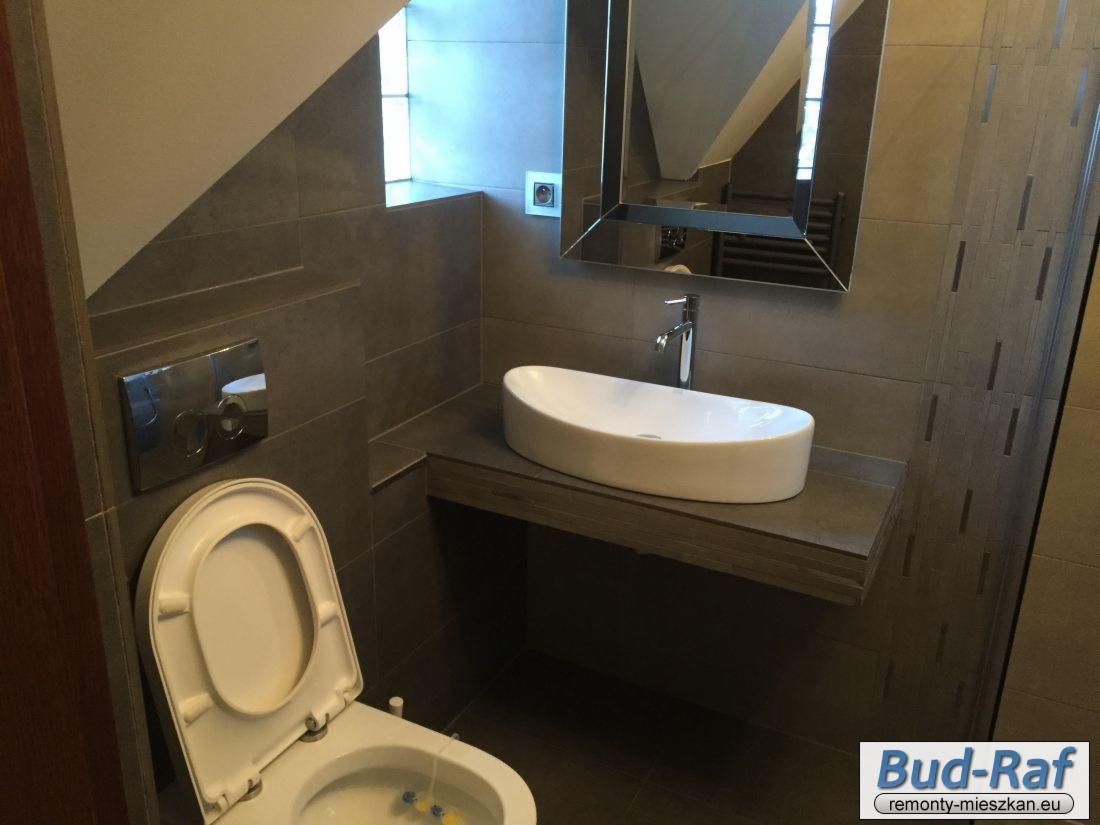 Nowoczesna łazienka z szarą glazurą, wisząca umywalka na drewnianym blacie i podwieszana toaleta z chromowanym przyciskiem spłukiwania, widoczne lustro w geometrycznej ramie.