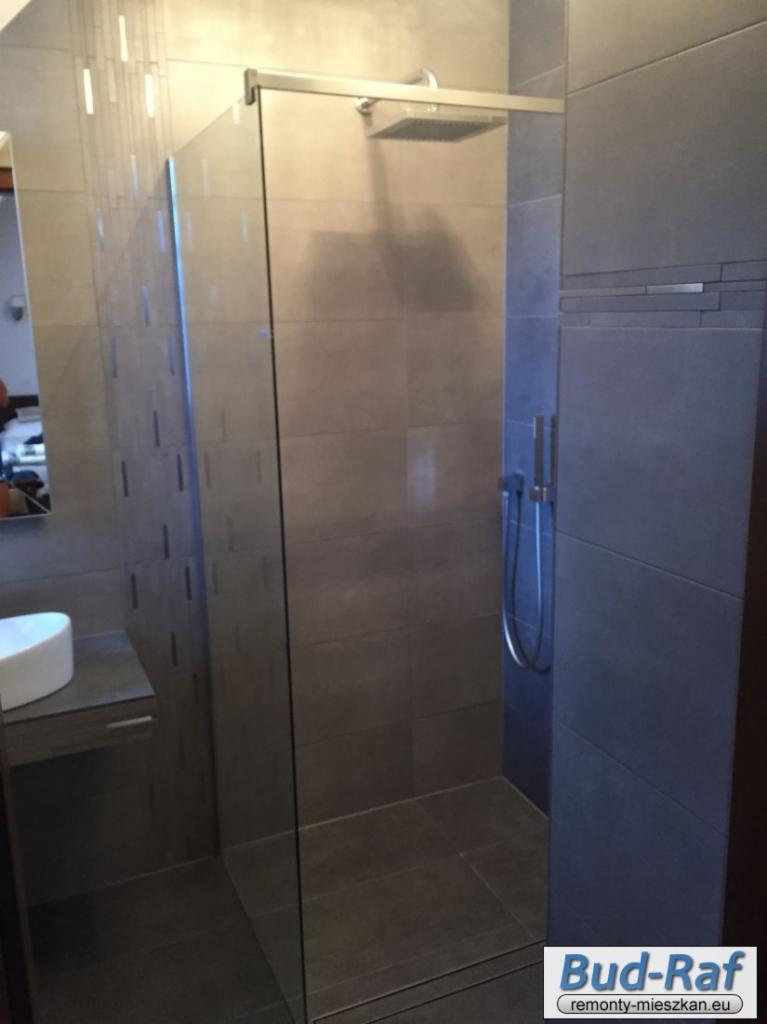 Nowoczesna kabina prysznicowa z szarą glazurą, deszczownicą i szklaną ścianą, widoczna umywalka na blacie, oświetlenie punktowe sufitowe.