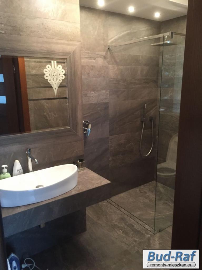 Nowoczesna łazienka z szarymi płytkami imitującymi marmur, owalna biała umywalka na kamiennym blacie i kabina prysznicowa ze szklanymi drzwiami.