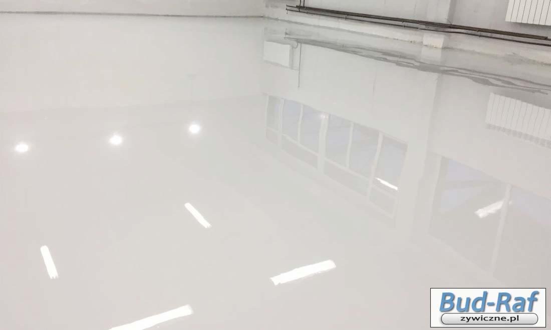 Błyszcząca, biała, epoksydowa posadzka przemysłowa z widocznymi odbiciami okien i oświetlenia sufitowego w hali produkcyjnej, z logo firmy w prawym dolnym rogu.