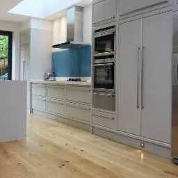 Nowoczesna kuchnia z wyspą, drewnianą podłogą i dużym oknem na ogród, oświetlona naturalnym światłem wpadającym przez świetlik.