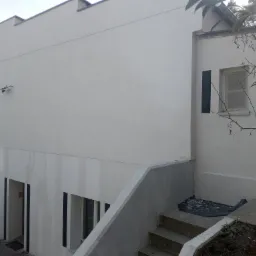 Elewacja budynku po malowaniu na biało, widoczne schody wejściowe i fragment ogrodu. Minimalistyczny styl, czarne obramowania okien.