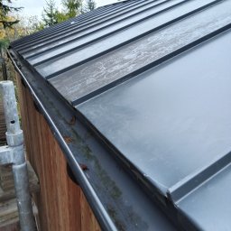 Bestdekarz Sp. z o.o. - Szary dach z blachy trapezowej z rynną, widok z dołu pod kątem. Na dachu widoczne ślady po deszczu. Obok dachu widoczna drewniana ściana budynku i fragment rusztowania.