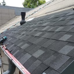 Bestdekarz Sp. z o.o. - Krycie dachu gontem bitumicznym w Gdańsku. Widoczny fragment dachu z kominkiem wentylacyjnym i narzędzia dekarskie. Częściowo widoczna membrana dachowa.