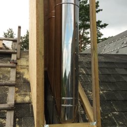 Bestdekarz Sp. z o.o. - Montaż komina systemowego na dachu pokrytym papą. Widoczna drewniana konstrukcja wspierająca i elementy blacharskie. Prace dekarskie w toku.