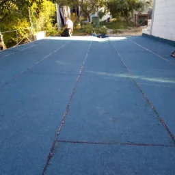 Niebieska, płaska powierzchnia dachu pokryta papą w Gdańsku, z widocznymi łączeniami i pracującym dekarzem w tle.