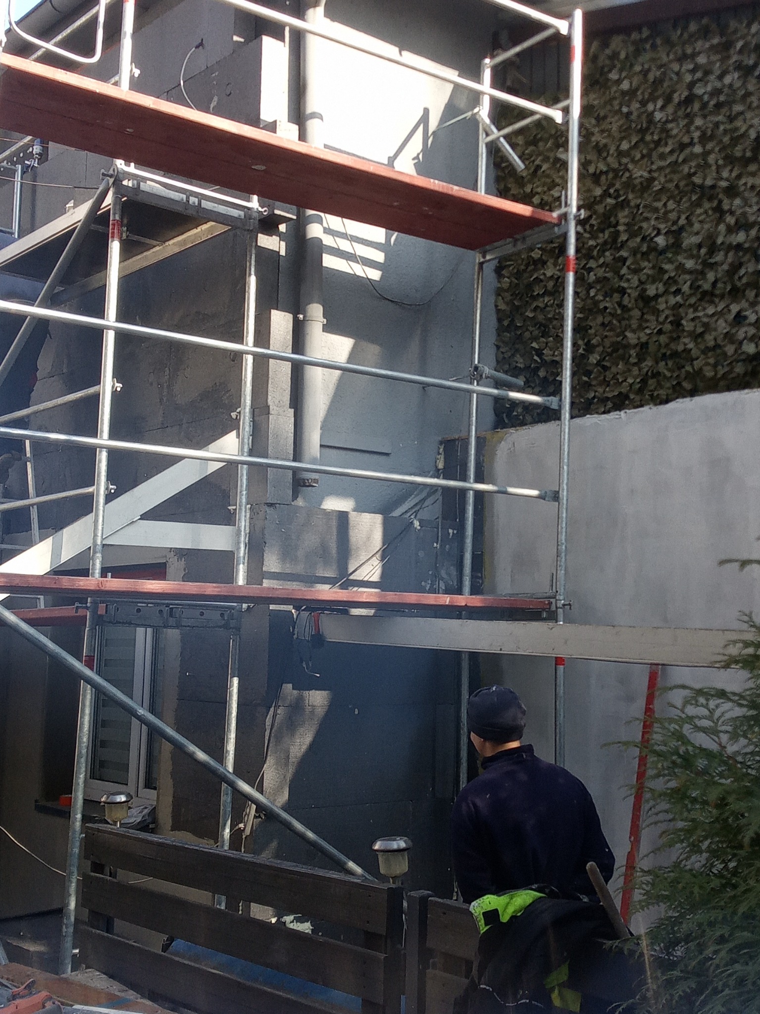 Elewacja budynku w trakcie ocieplania, widoczne rusztowania, fragment elewacji z ociepleniem i siatką, obok fragment ściany z zieloną okładziną.