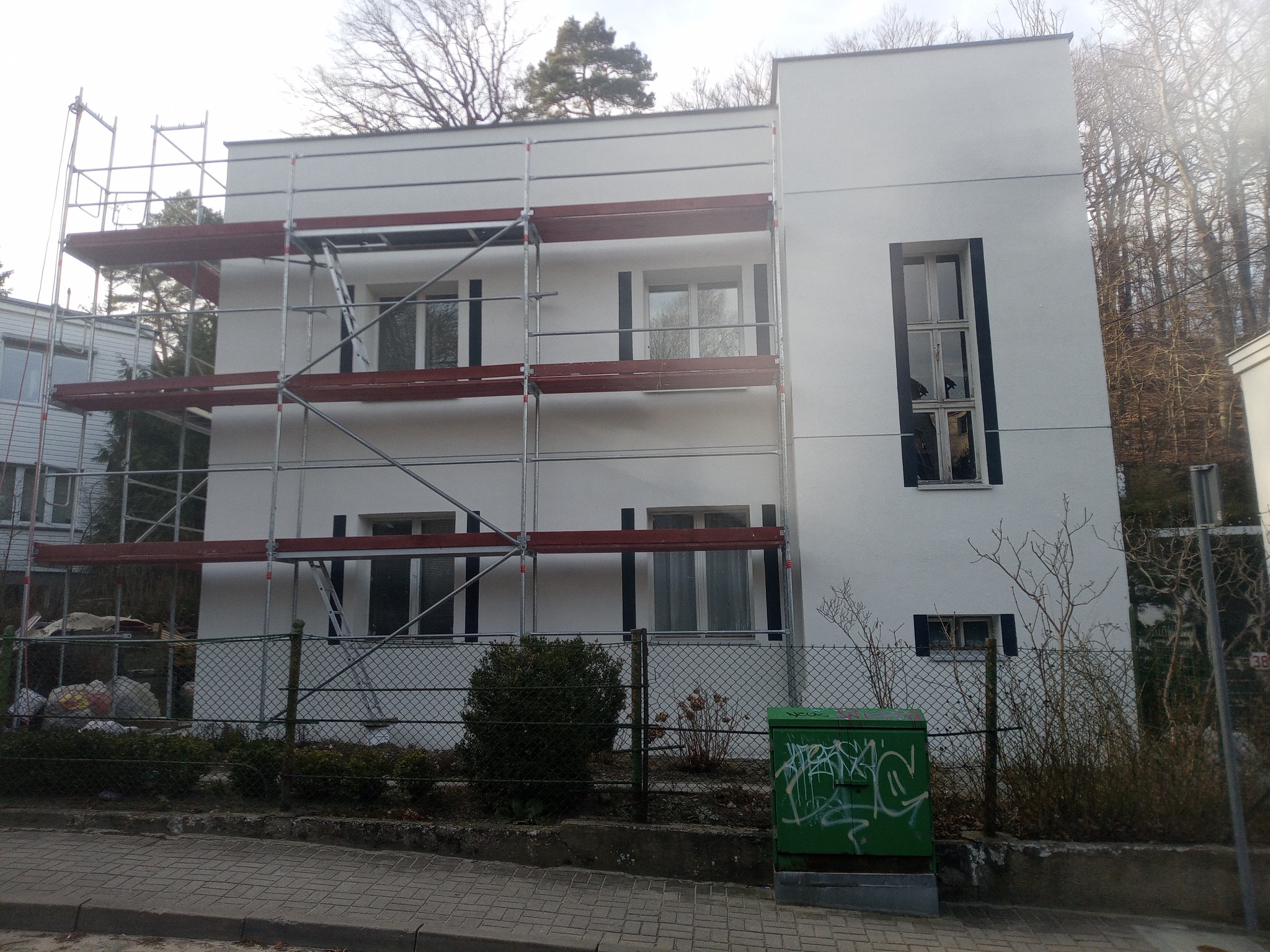Biały budynek z rusztowaniem w trakcie prac elewacyjnych, widoczne okna z czarnymi ramami i zielona skrzynka elektryczna z graffiti na pierwszym planie.