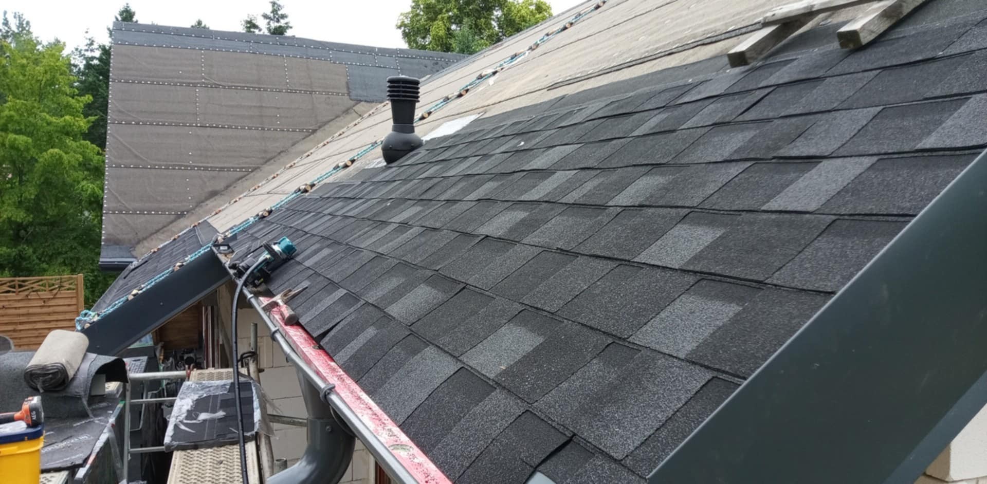 Krycie dachu gontem bitumicznym w Gdańsku. Widoczny fragment dachu z kominkiem wentylacyjnym i narzędzia dekarskie. Częściowo widoczna membrana dachowa.