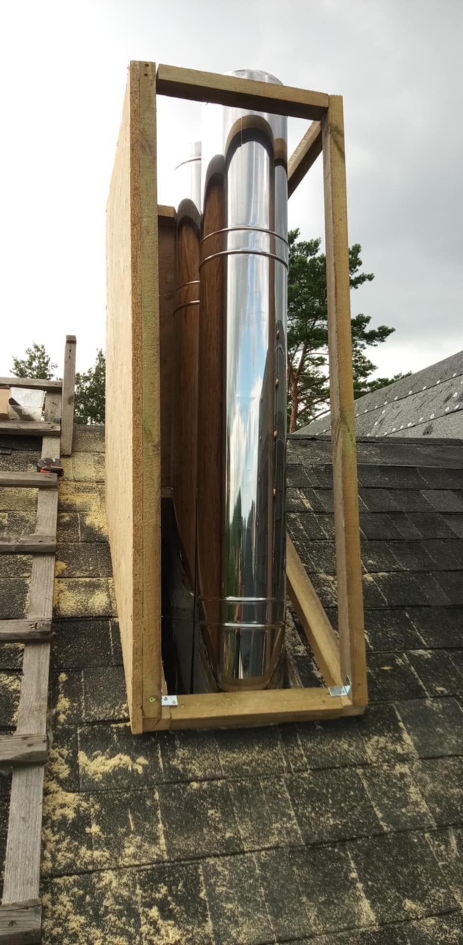 Montaż komina systemowego na dachu pokrytym papą. Widoczna drewniana konstrukcja wspierająca i elementy blacharskie. Prace dekarskie w toku.