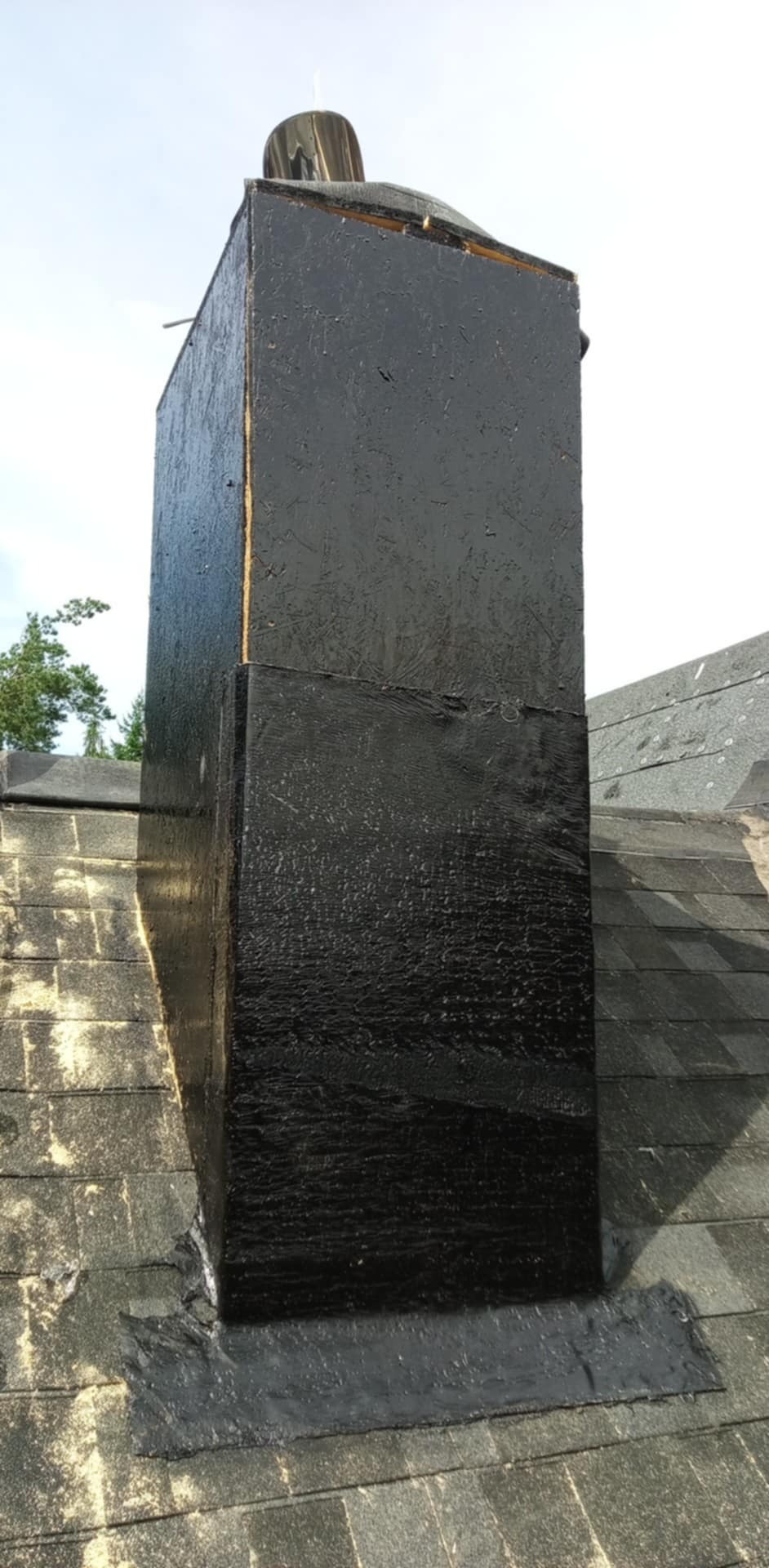 Czarny, zabezpieczony komin na dachu z szarych dachówek. Widoczne ślady uszczelniania i zabezpieczania podstawy komina. Komin pomalowany na czarno.
