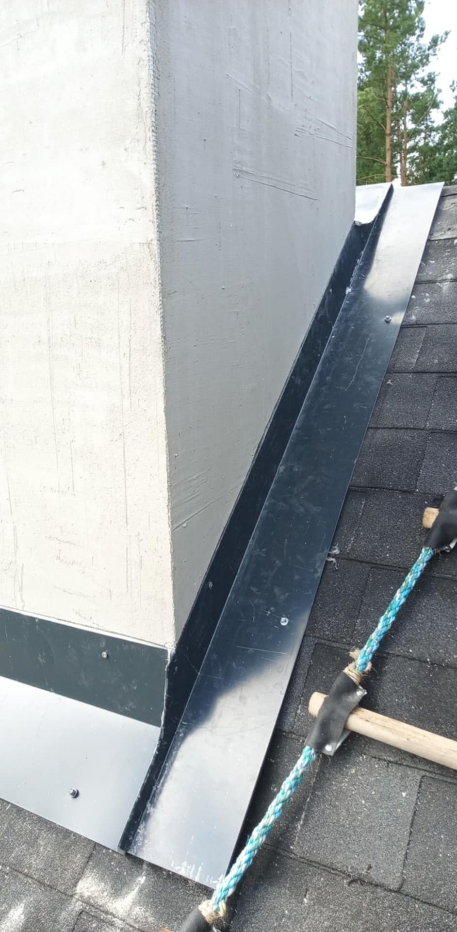 Szczegółowe ujęcie obróbki blacharskiej komina na dachu pokrytym gontem bitumicznym, z widoczną liną zabezpieczającą i fragmentem lasu w tle.