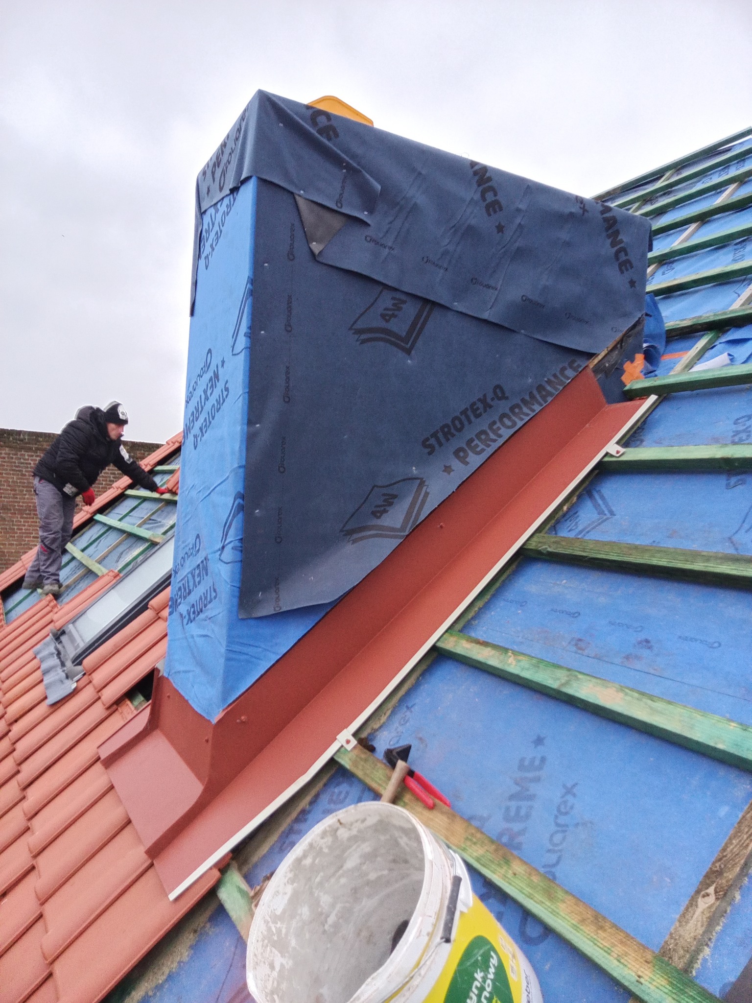 Montaż obróbki blacharskiej komina na dachu pokrytym czerwoną dachówką. Widoczna membrana dachowa i drewniana konstrukcja. Pracownik w trakcie pracy.