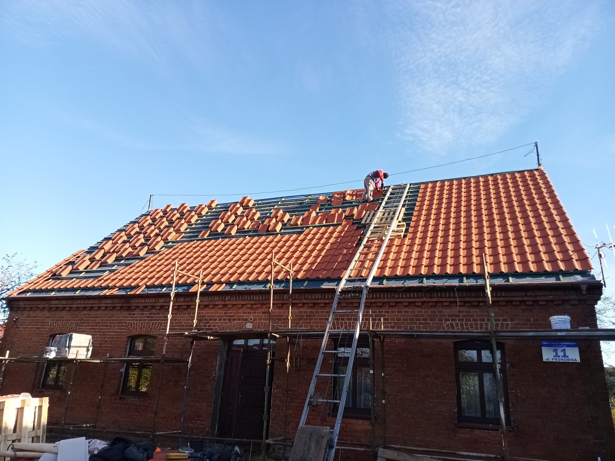 Ceglany dom z częściowo wymienionym dachem z czerwonej dachówki. Na dachu drabina i dekarz w trakcie pracy. Widoczne rusztowania i tabliczka z adresem.