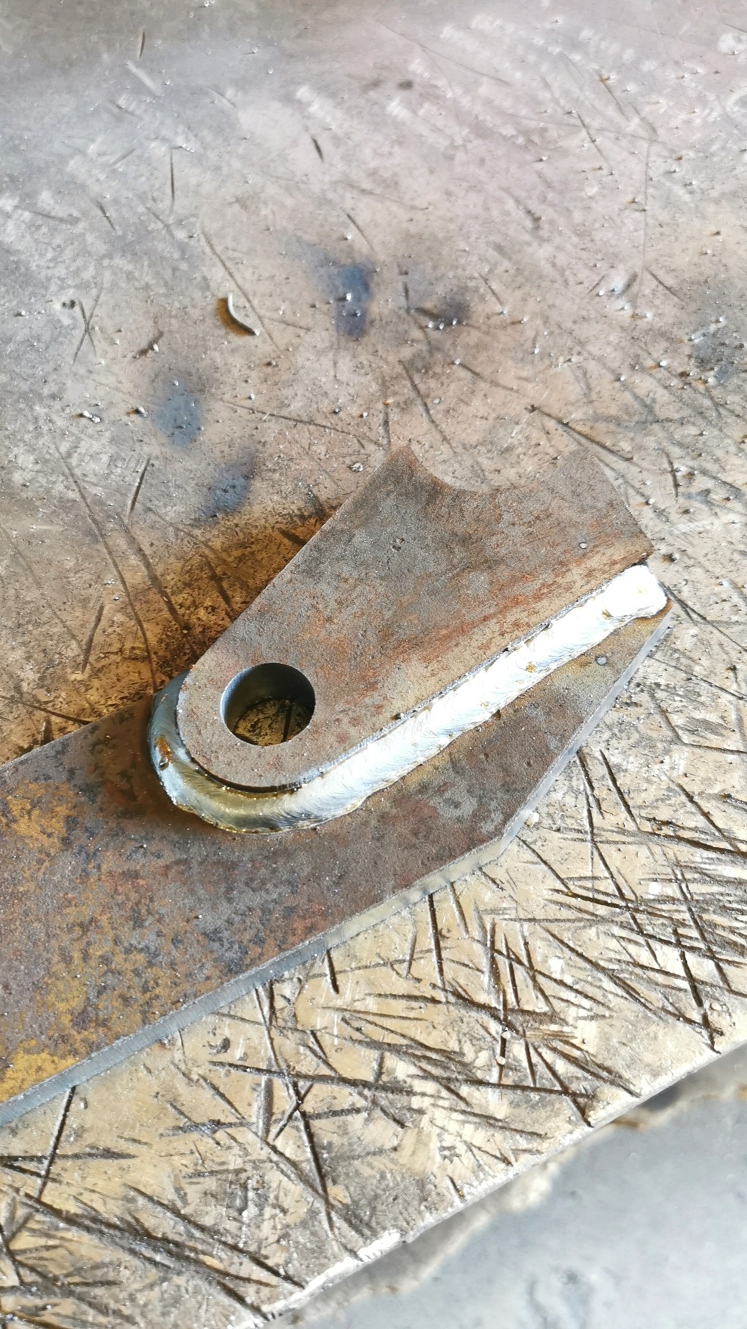 Dwa kawałki zardzewiałego metalu połączone świeżym, jasnym spawem na szorstkim, porysowanym stole warsztatowym. Jeden element ma okrągły otwór.