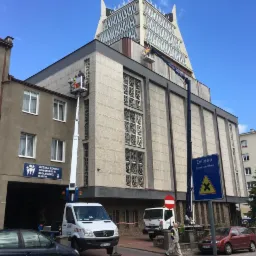 Dwóch pracowników na podnośnikach koszowych, wykonujących prace na elewacji wysokiego, modernistycznego budynku z elementami ażurowymi, w tle błękitne niebo i inne budynki.