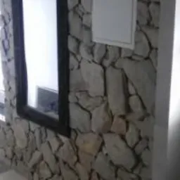 Ściana wewnętrzna wyłożona nieregularnymi kamieniami w odcieniach szarości i beżu, z widocznym oknem z czarną ramą i białym parapetem. Na ścianie widoczna biała skrzynka inspekcyjna. Ujęcie...