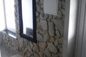 Ściana wewnętrzna wyłożona nieregularnymi kamieniami w odcieniach szarości i beżu, z widocznym oknem z czarną ramą i białym parapetem. Na ścianie widoczna biała skrzynka inspekcyjna. Ujęcie...