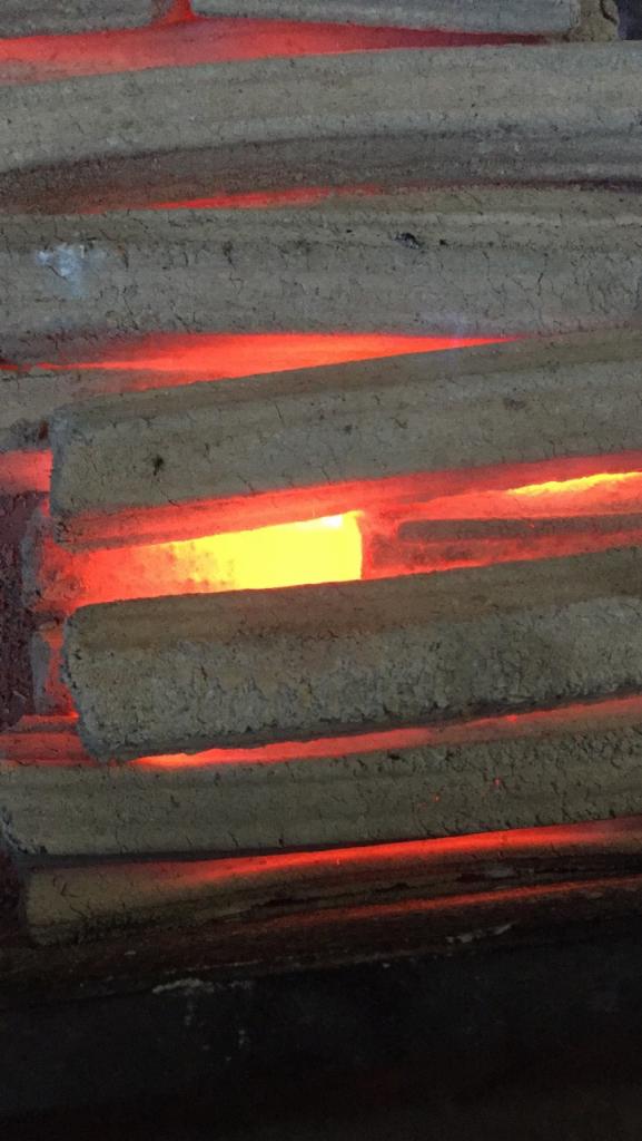 Ułożone obok siebie, prostokątne brykiety z węgla drzewnego Pini Kay, pomiędzy którymi żarzy się pomarańczowo-żółty ogień.