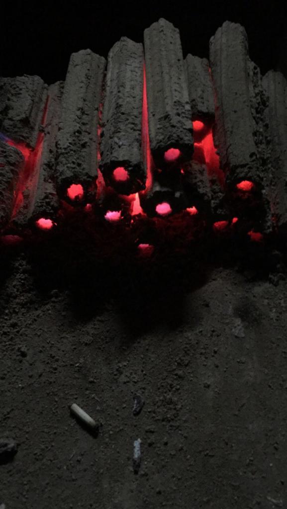 Płonące brykiety Pini Kay w ciemności, widoczne żarzące się otwory i niedopałek na ziemi.