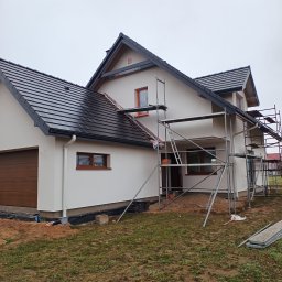 BudiLuk Łukasz Żdanowicz - Elewacja domu jednorodzinnego w trakcie prac wykończeniowych. Widoczne rusztowania, jasna elewacja, ciemna dachówka i brązowe okna. Pochmurne niebo w tle.