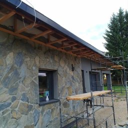 BudiLuk Łukasz Żdanowicz - Elewacja domu w trakcie wykończenia z kamienia naturalnego. Widoczne rusztowania i drewniana konstrukcja dachu. Okno z butelką płynu do czyszczenia.