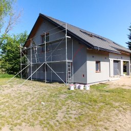 BudiLuk Łukasz Żdanowicz - Dom jednorodzinny w trakcie tynkowania elewacji, z widocznymi rusztowaniami i wiaderkami z farbą. Świeżo otynkowana szara elewacja w słoneczny dzień.