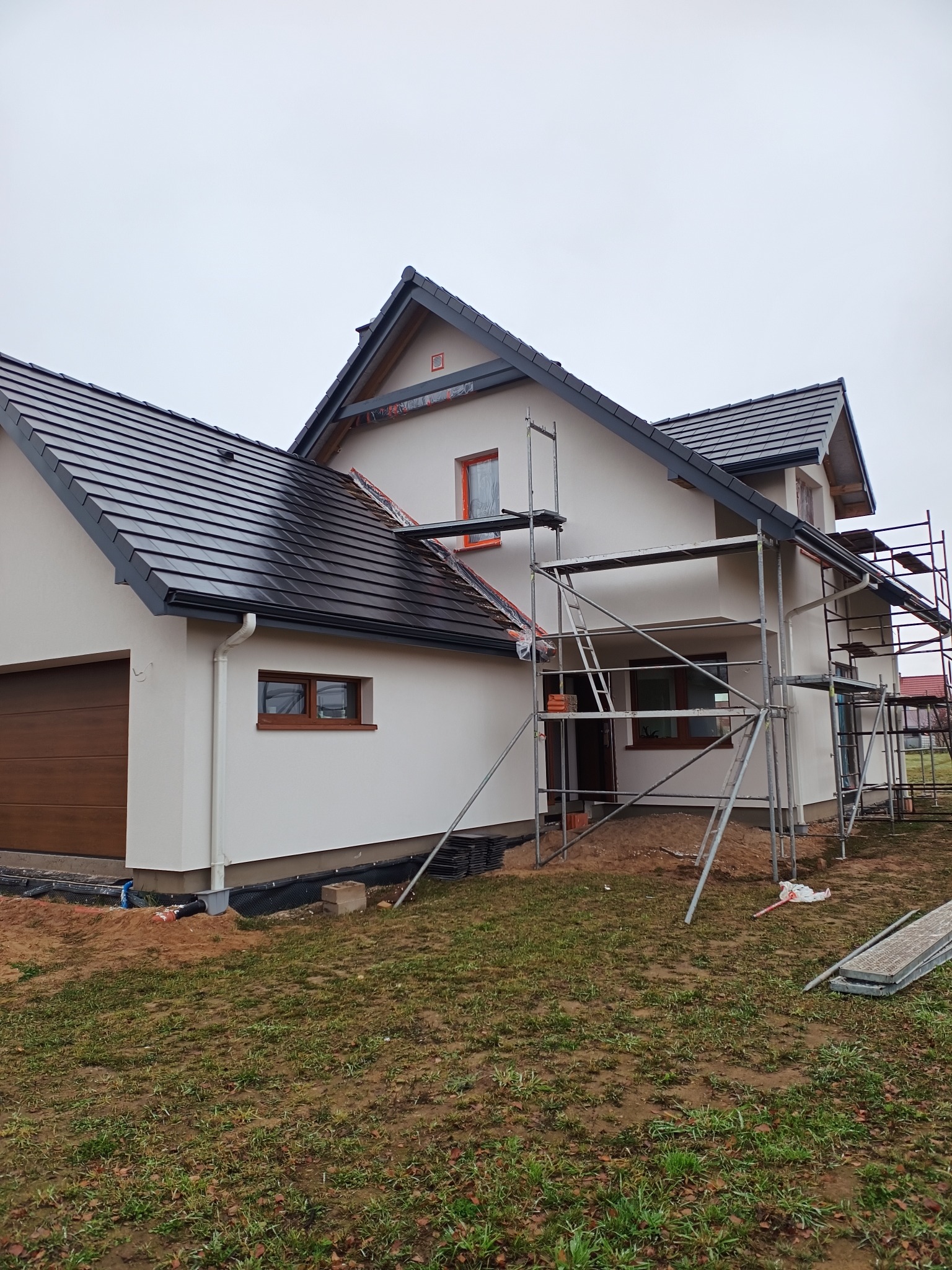 Elewacja domu jednorodzinnego w trakcie prac wykończeniowych. Widoczne rusztowania, jasna elewacja, ciemna dachówka i brązowe okna. Pochmurne niebo w tle.