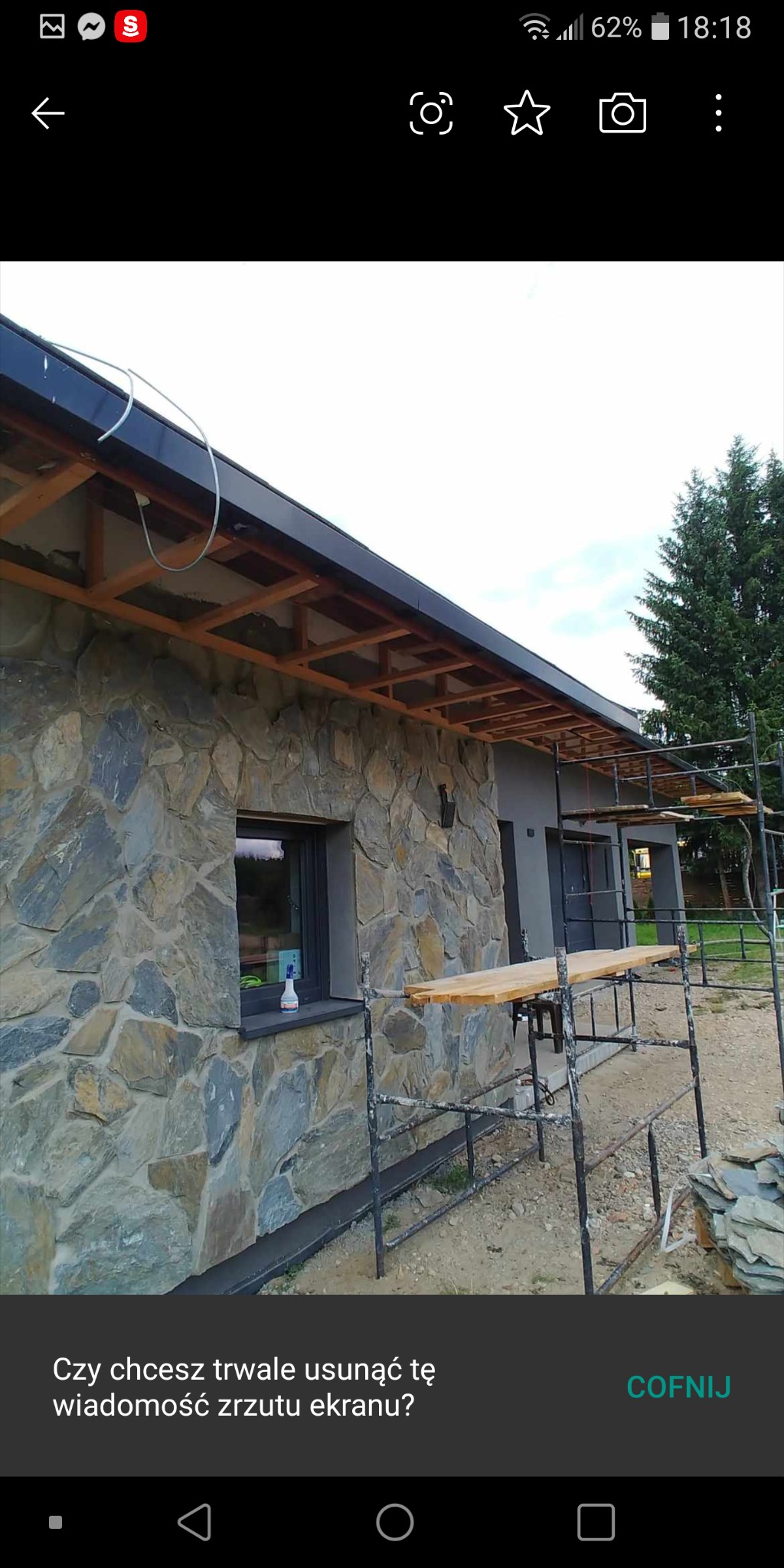 Elewacja domu w trakcie wykończenia z kamienia naturalnego. Widoczne rusztowania i drewniana konstrukcja dachu. Okno z butelką płynu do czyszczenia.