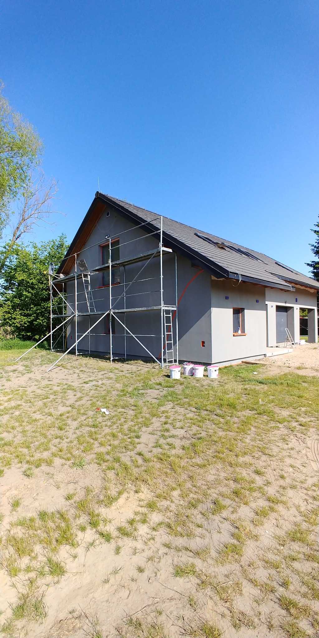 Dom jednorodzinny w trakcie tynkowania elewacji, z widocznymi rusztowaniami i wiaderkami z farbą. Świeżo otynkowana szara elewacja w słoneczny dzień.