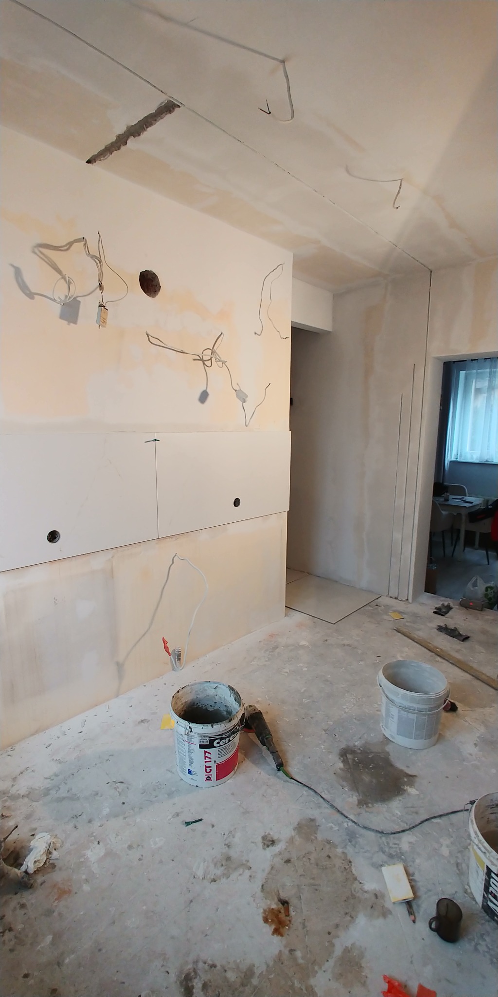 Wnętrze w trakcie prac wykończeniowych: ściany z płytami, widoczne przewody elektryczne, wiadra z zaprawą i szlifierka na podłodze. Etap przygotowania do gładzi.