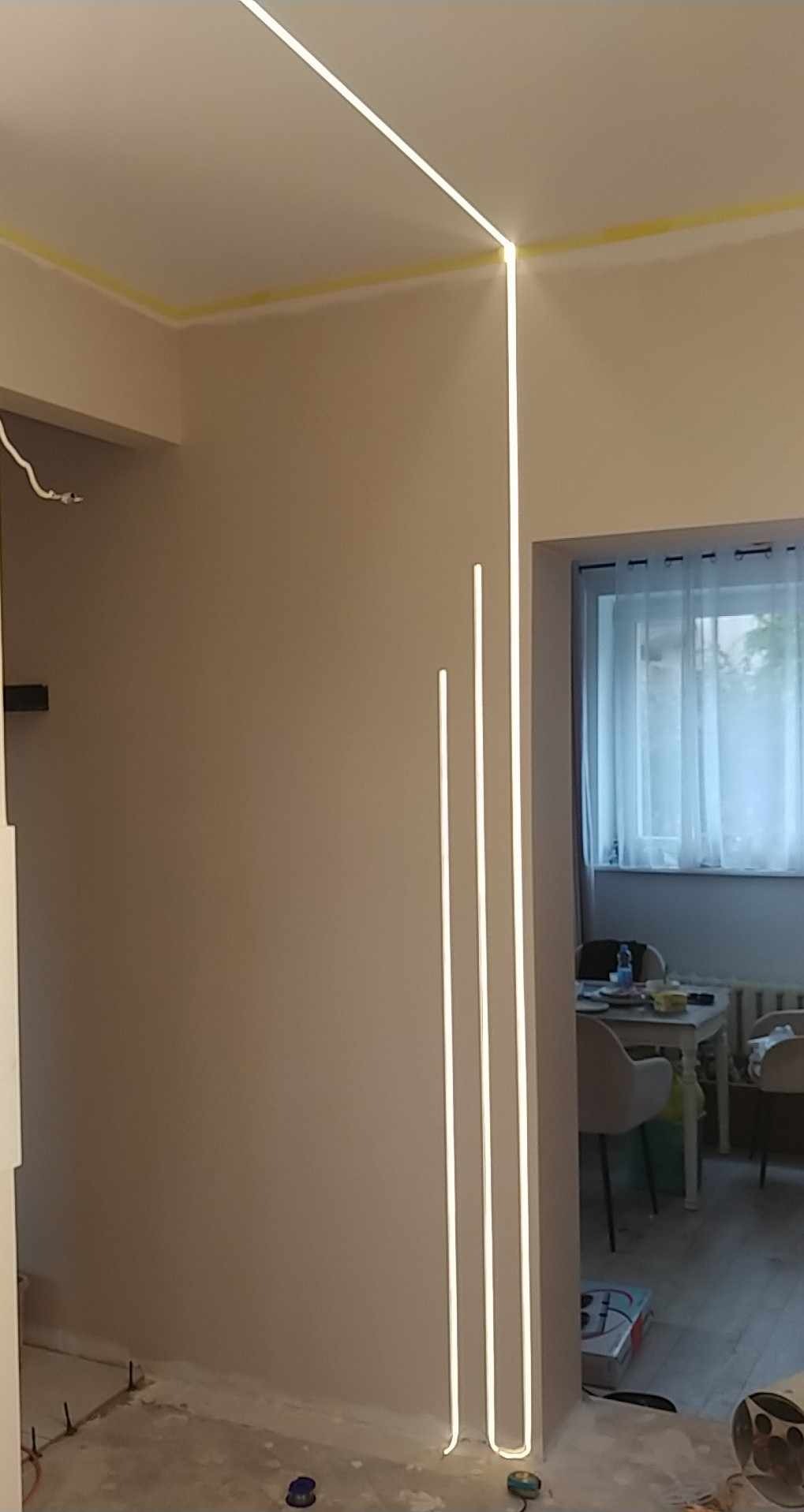 Nowoczesne oświetlenie LED w rogu pokoju z szarą ścianą. Pionowe i poziome linie świetlne tworzą minimalistyczny design. Żółta taśma malarska na suficie.