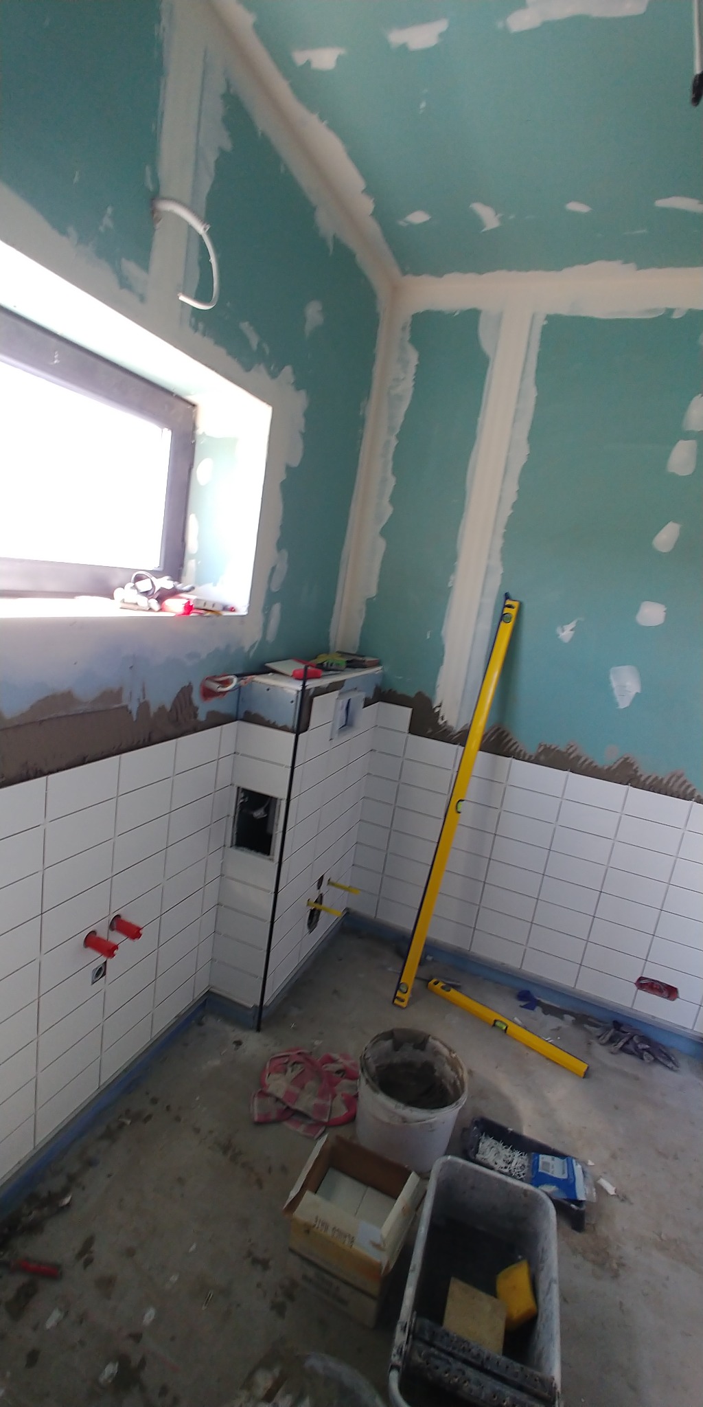Wnętrze łazienki w trakcie remontu: białe płytki na ścianach do połowy wysokości, surowy sufit z płyt karton-gips, poziomice i narzędzia na podłodze.