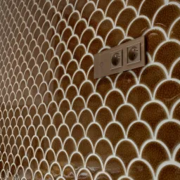 Ściana wyłożona błyszczącymi, brązowymi płytkami w kształcie rybiej łuski z białymi fugami, widoczne gniazdka elektryczne i wystające kable instalacji elektrycznej w trakcie montażu.