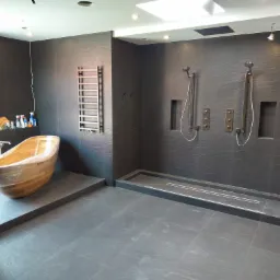 Nowoczesna łazienka w trakcie remontu z drewnianą wanną owiniętą folią ochronną, czarne płytki na ścianach i podłodze, oraz zamontowaną armaturą prysznicową i grzejnikiem drabinkowym.