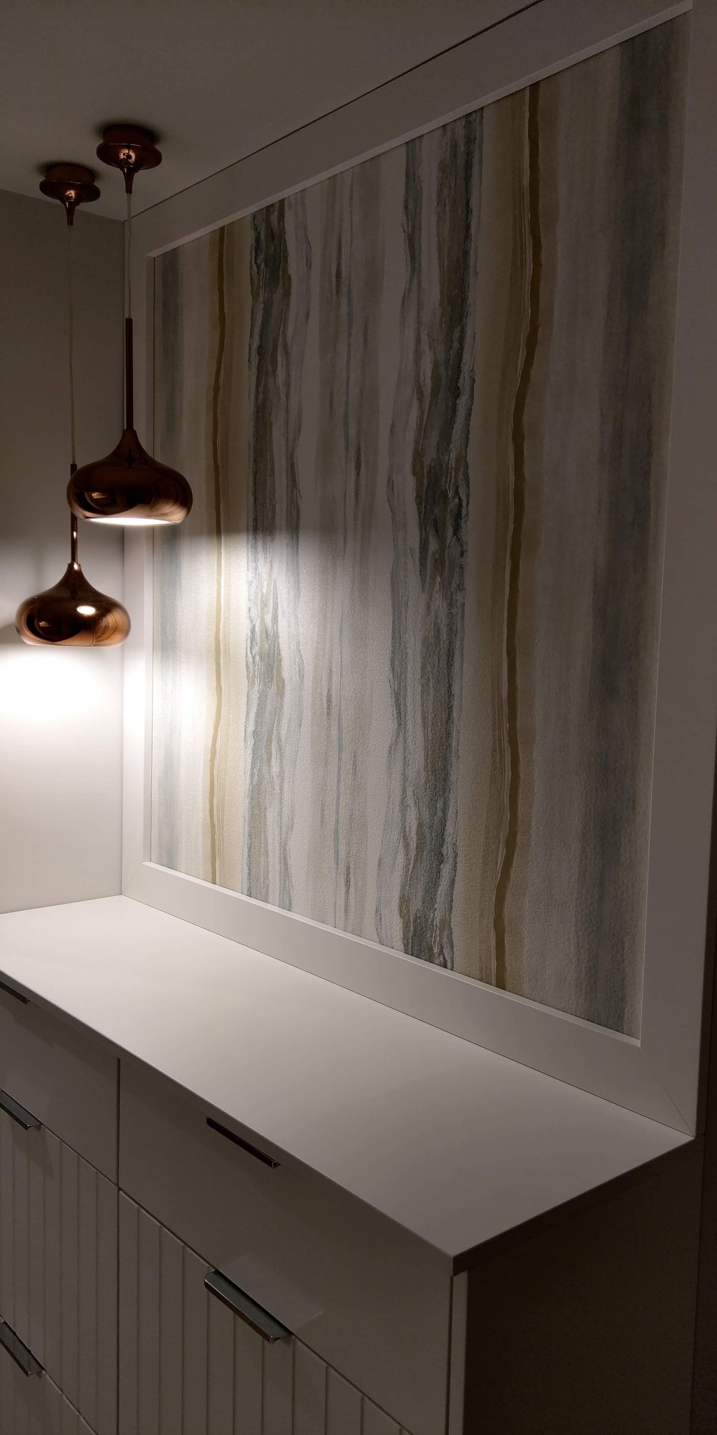 Nowoczesne wykończenie wnętrza z dekoracyjną tapetą w odcieniach szarości i beżu, oświetloną przez dwie miedziane lampy wiszące nad białą szafką z szufladami.