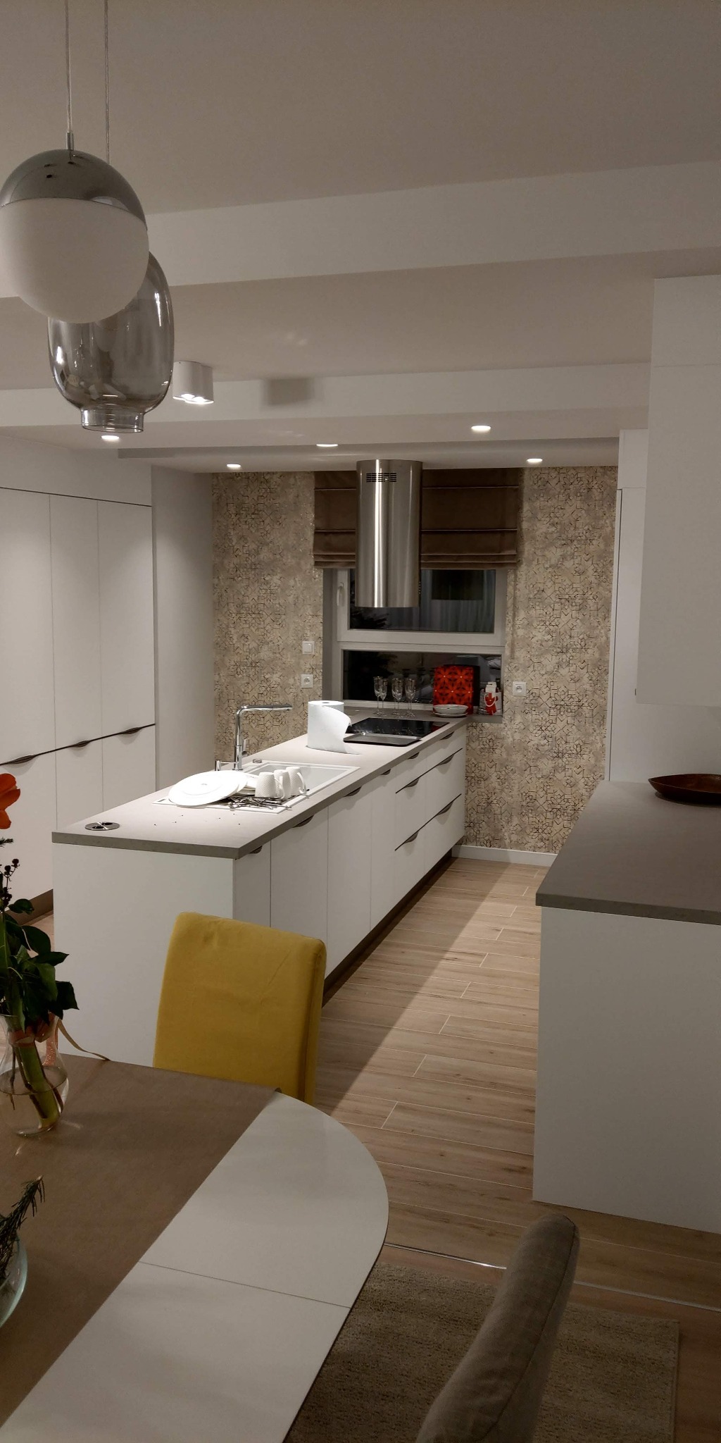 Nowoczesna kuchnia z wyspą, białymi szafkami i drewnopodobną podłogą; na blacie wyspy naczynia w zlewie, nad wyspą srebrny okap, w tle tapeta o geometrycznym wzorze i okno z brązowymi roletami...