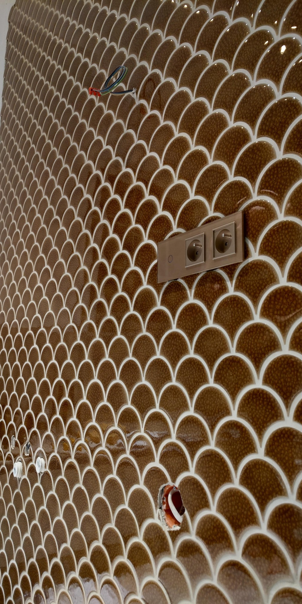 Ściana wyłożona błyszczącymi, brązowymi płytkami w kształcie rybiej łuski z białymi fugami, widoczne gniazdka elektryczne i wystające kable instalacji elektrycznej w trakcie montażu.