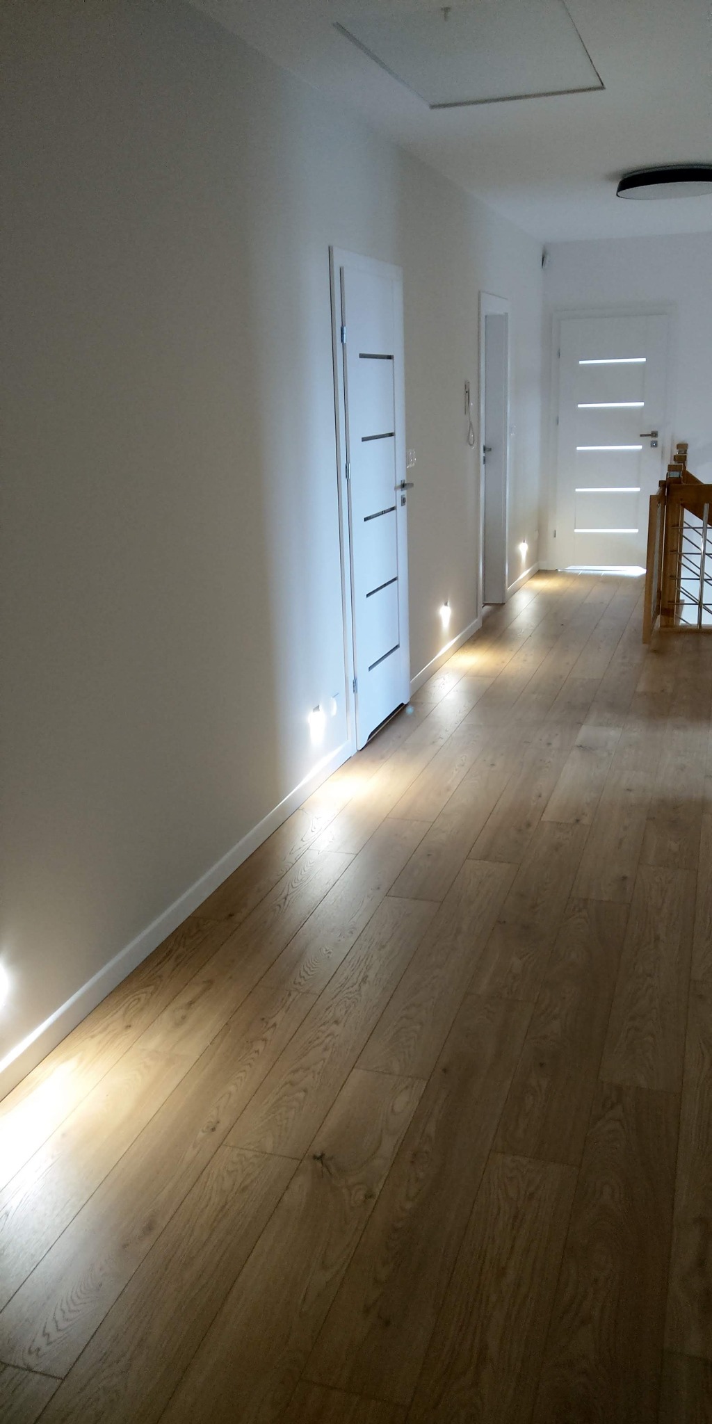 Korytarz z drewnianą podłogą oświetloną punktowymi lampami podłogowymi, białe ściany i drzwi z poziomymi wstawkami, widoczne wejście na strych w suficie.