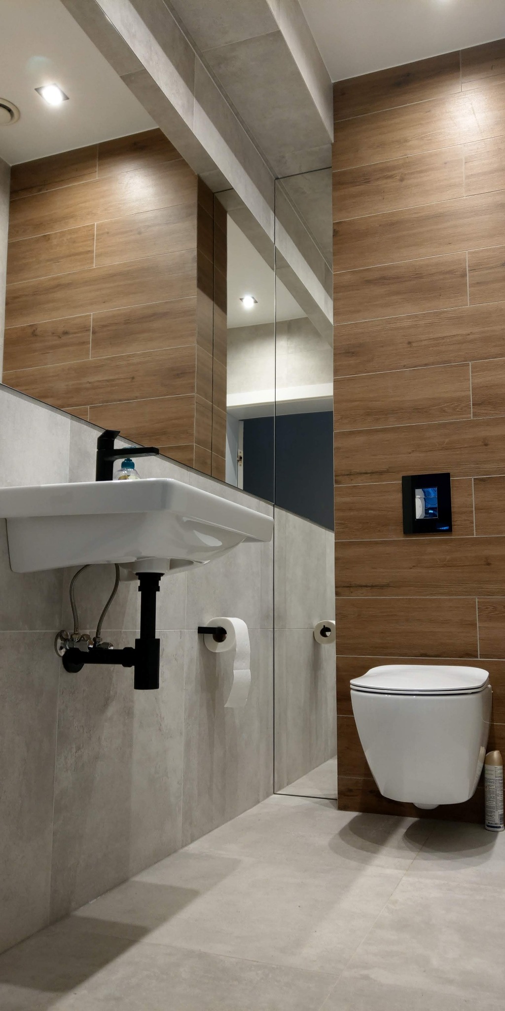 Nowoczesna łazienka z drewnopodobnymi płytkami, podwieszaną toaletą i umywalką z czarną armaturą, minimalistyczny design.