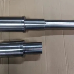 Dwa metalowe wałki o różnych rozmiarach, obrobione z widocznymi śladami obróbki, leżące na tekturowym tle.