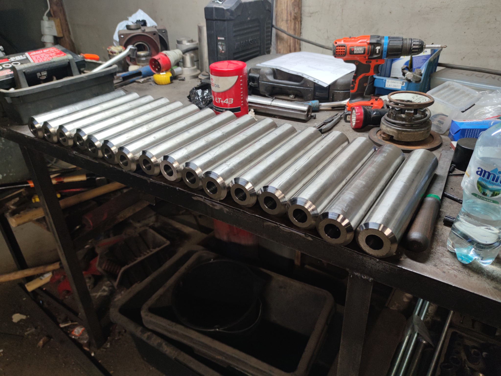 Rząd obrobionych, metalowych cylindrów z otworem, leżących na warsztatowym stole, widoczne narzędzia i akcesoria, w tle ściana warsztatu.